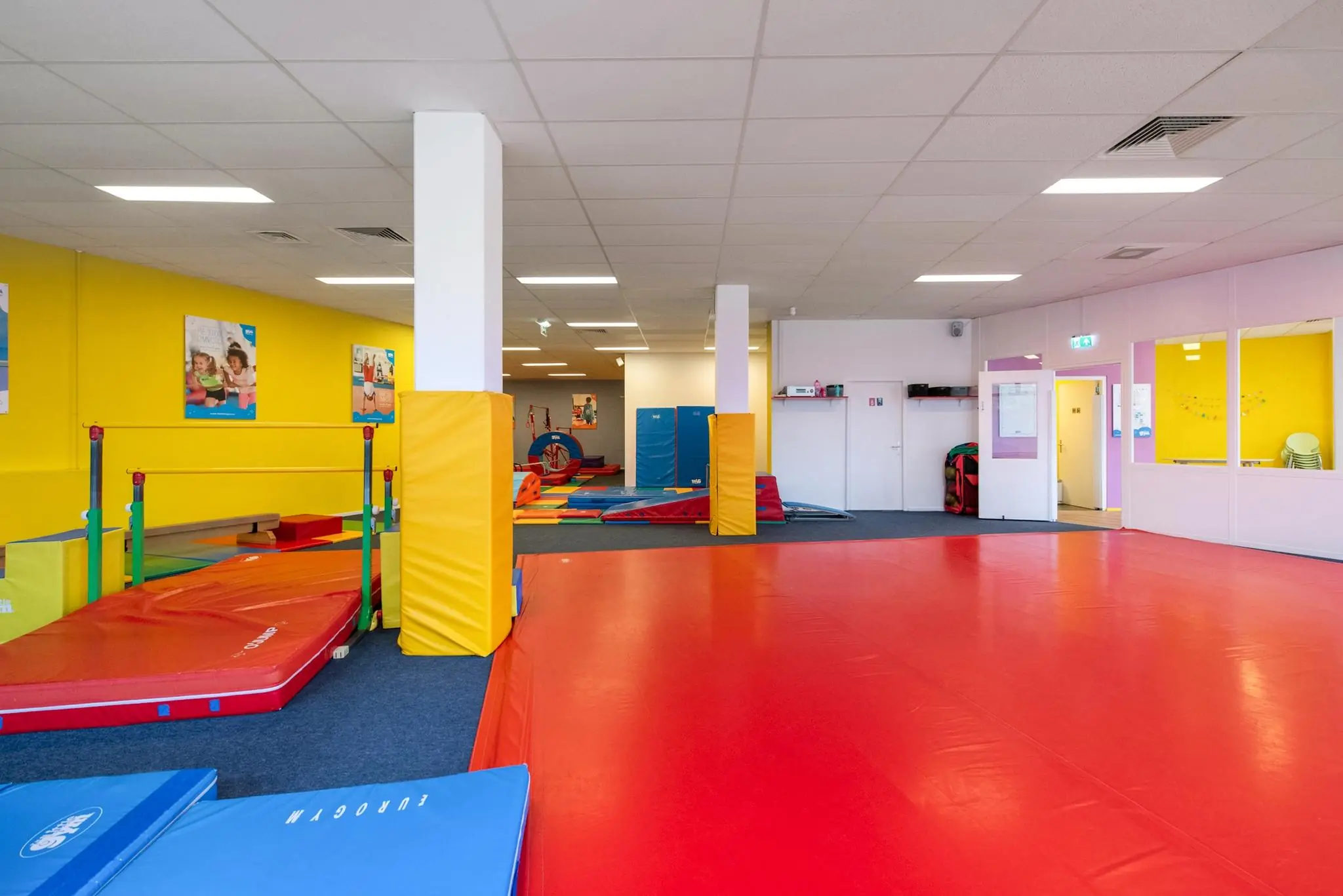 Kleurrijke binnenruimte van een kindergymzaal aan de Koningin Julianalaan met matten, turntoestellen en een gele muur.